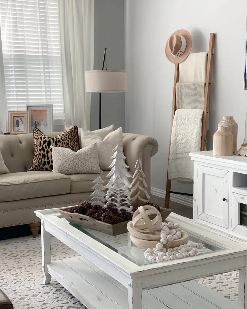 A neutral-toned living room with a tufted sofa, neutral patterned pillows, a glass coffee table with decorative objects, a floor lamp, and a blanket ladder by the wall styled for winter.