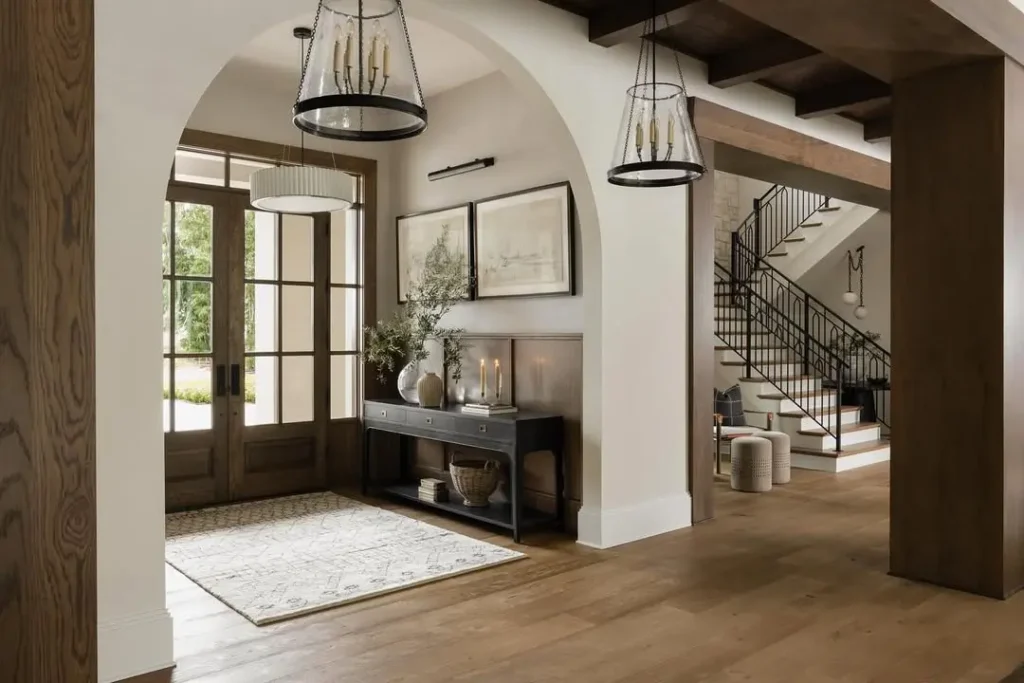 Spacious Organic Modern Entryway with wooden double doors, a console table with decor, large rug, arched entryway, and view of staircase with black railing in the background.