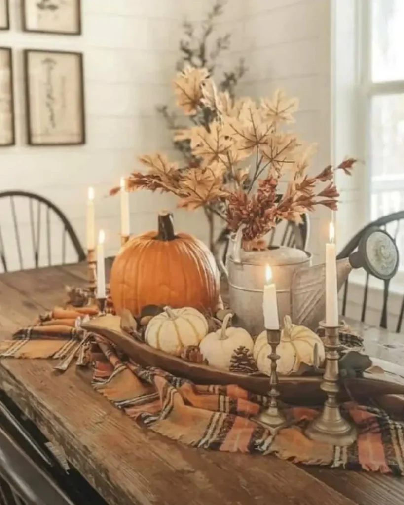 A wooden table decorated with pumpkins, candles, a plaid cloth, and a metal pitcher with autumn leaves creates a cozy fall-themed centerpiece.