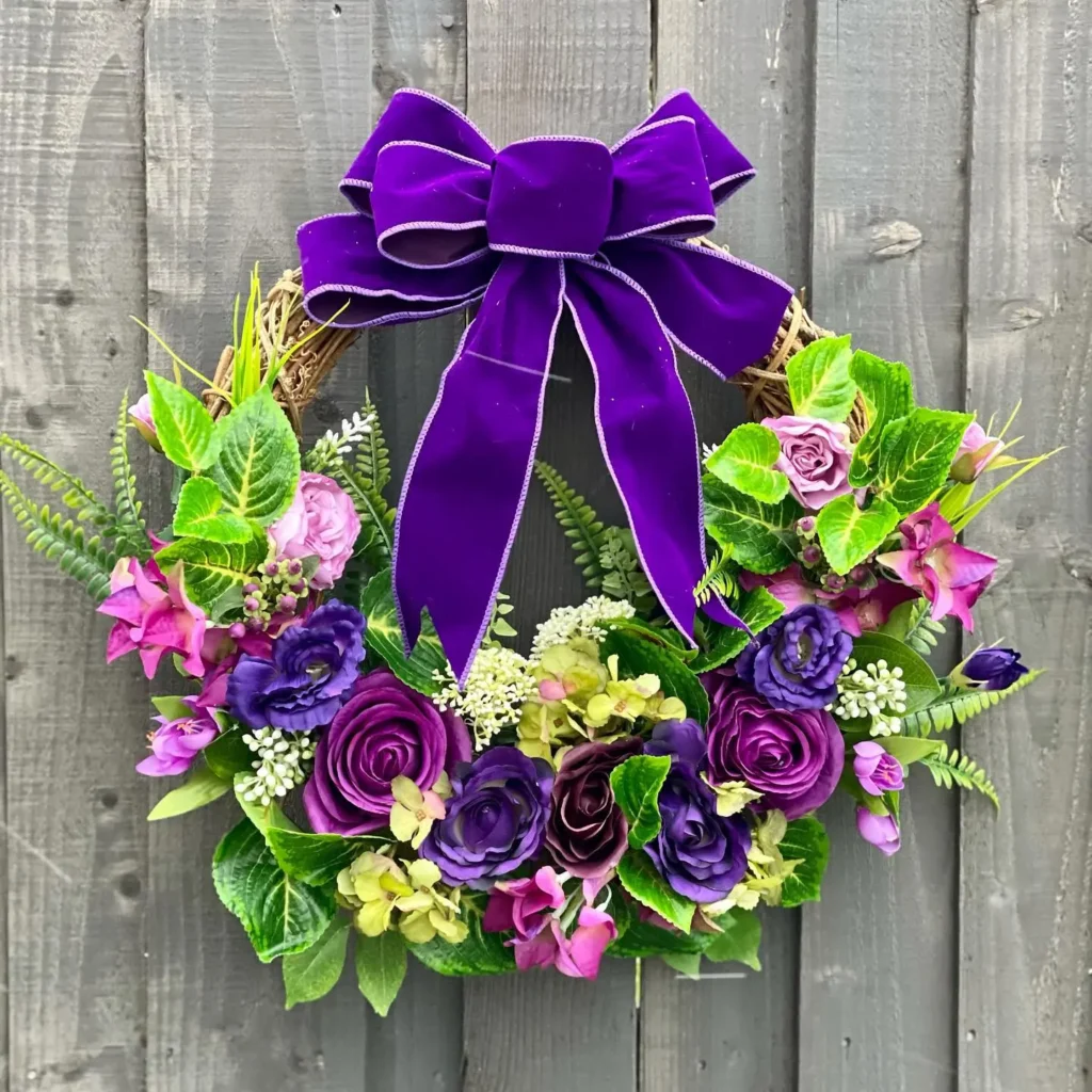 A decorative winter wreath with purple and violet flowers, green leaves, and a large purple ribbon bow, hanging on a wooden fence.