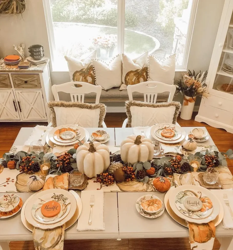 A dining table set for a fall-themed meal with white chairs, pumpkin and foliage centerpieces, and plates with "Thankful" written on them. Light streams in from a large window.
