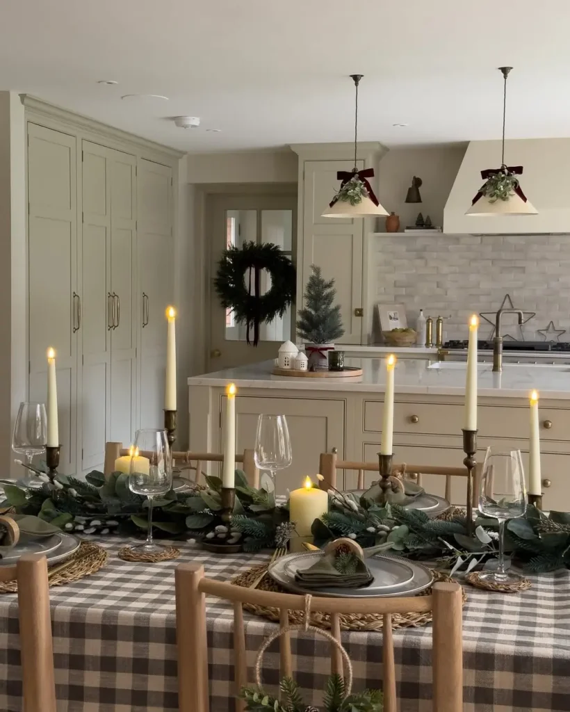 A rustic farmhouse Christmas dining table with a checkered tablecloth, candles, and greenery is set for a meal in a kitchen adorned with wreaths and festive accents.
