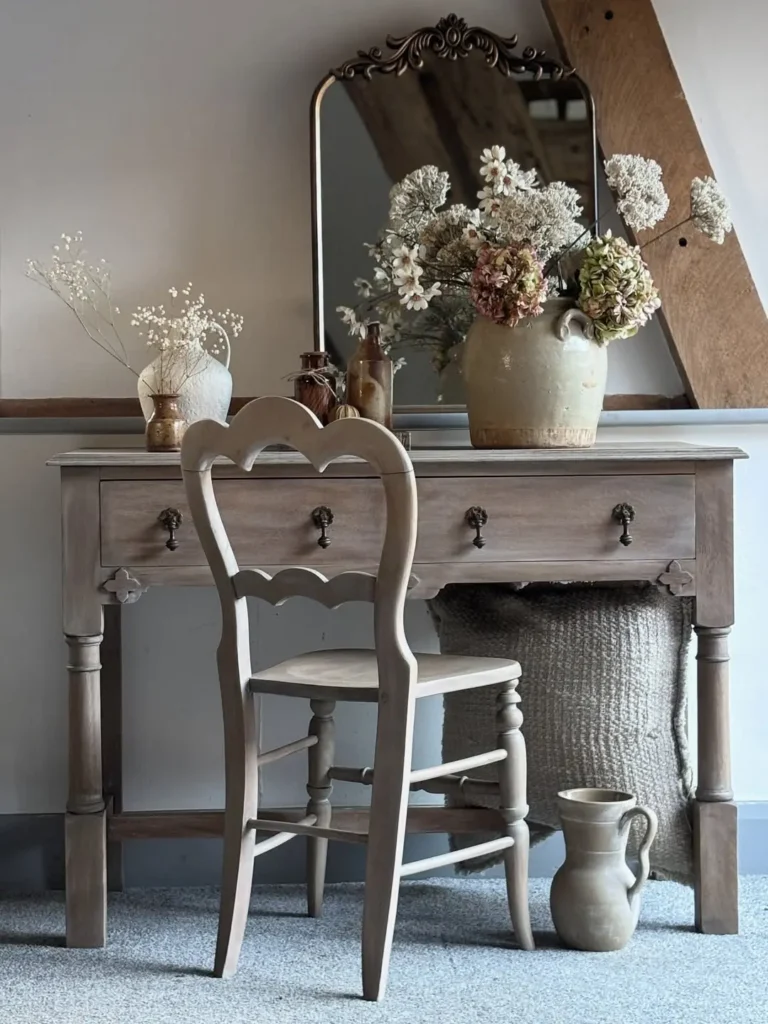 A wooden vanity table with a mirror, decorative vases of dried flowers, a matching chair, and a ceramic jug on a gray carpeted floor, perfect for elevating your space with charming rustic bedroom decor finds.