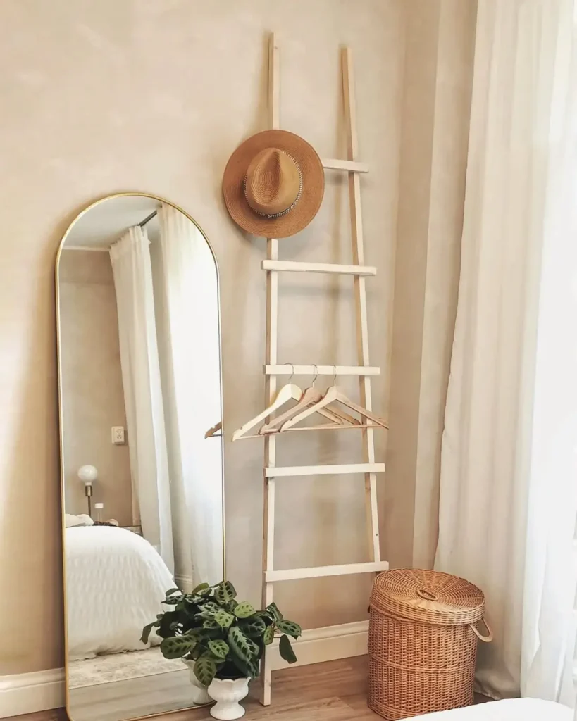 A wooden ladder with hangers and a straw hat leans against a wall beside a tall mirror, a rattan basket, a potted plant, and white curtains, perfect affordable rustic bedroom decor finds.