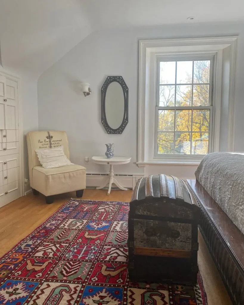 A bedroom with a large window, white walls, an upholstered chair, a small round table with a vase, a decorative mirror, a patterned rug, and rustic wooden trunk bedroom styling at the foot of the bed.
