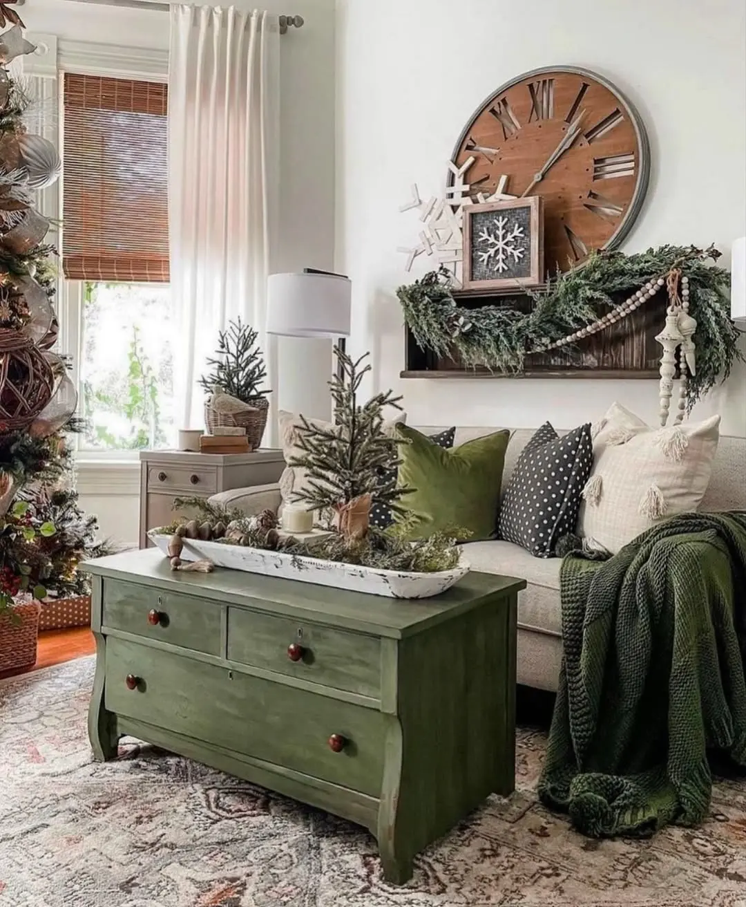 A cozy living room features a green chest as a coffee table, a sofa with cushions and blankets, holiday decorations, and a large wall clock above greenery, showcasing tips for decorating with texture and layer during winter months.