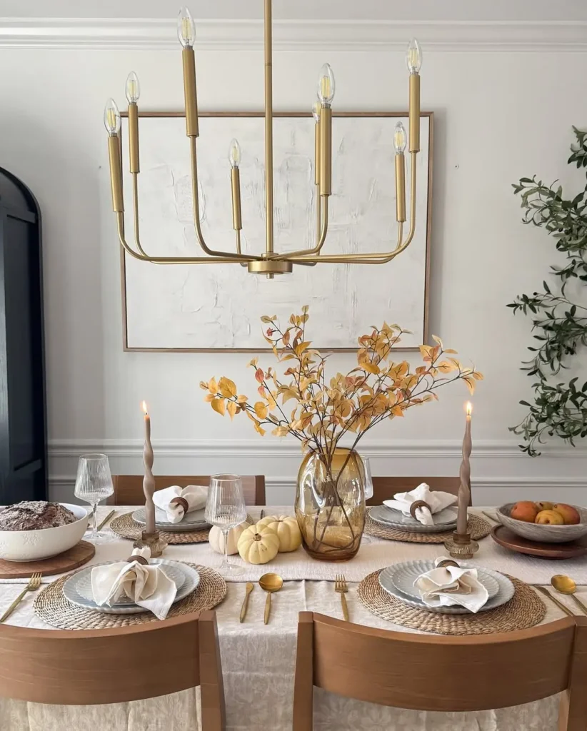 A minimalist Thanksgiving table set for four with neutral-toned plates, gold cutlery, candles, a glass vase with yellow leaves, pumpkins, and bowls of fruit under a gold chandelier.