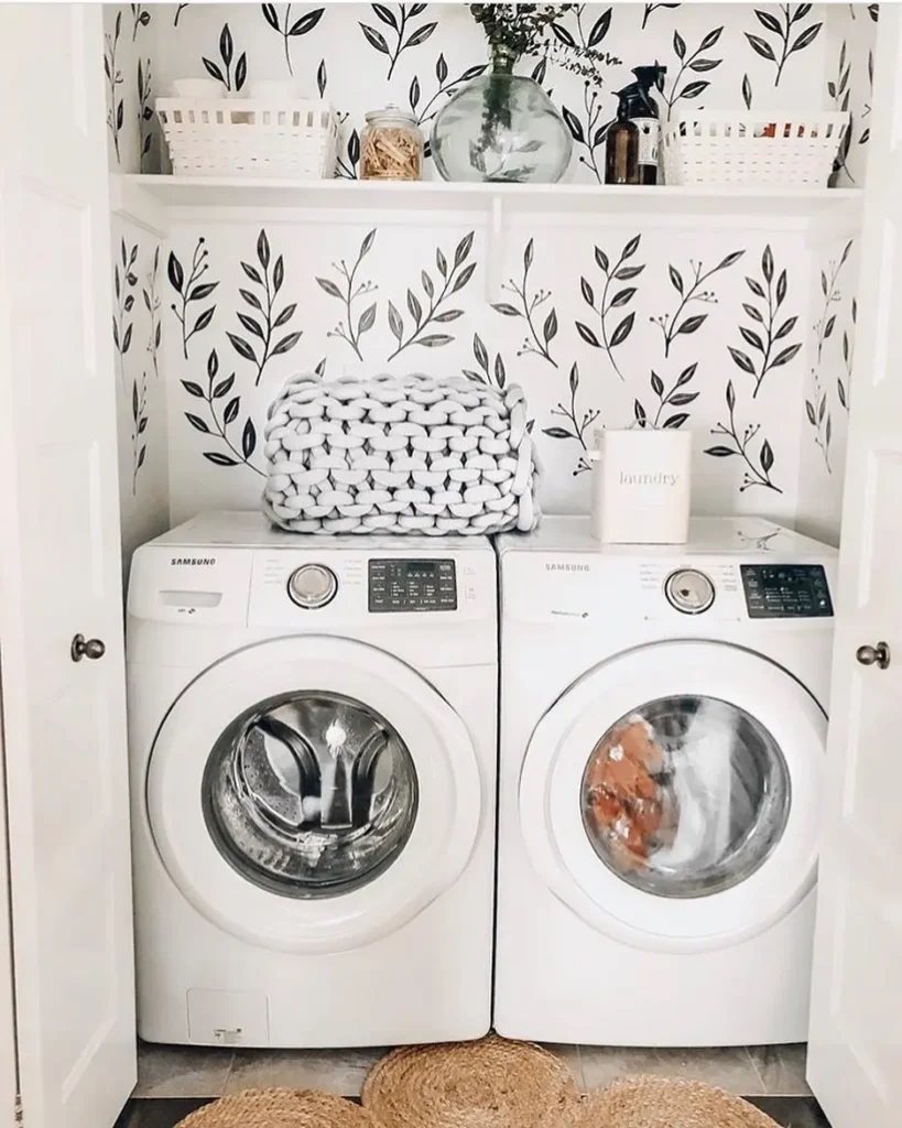 A small laundry closet with two front-loading washing machines, open shelves above hold baskets and provide storage space for towels, detergent, and a vase, all set against stylish leaf-patterned wallpaper.