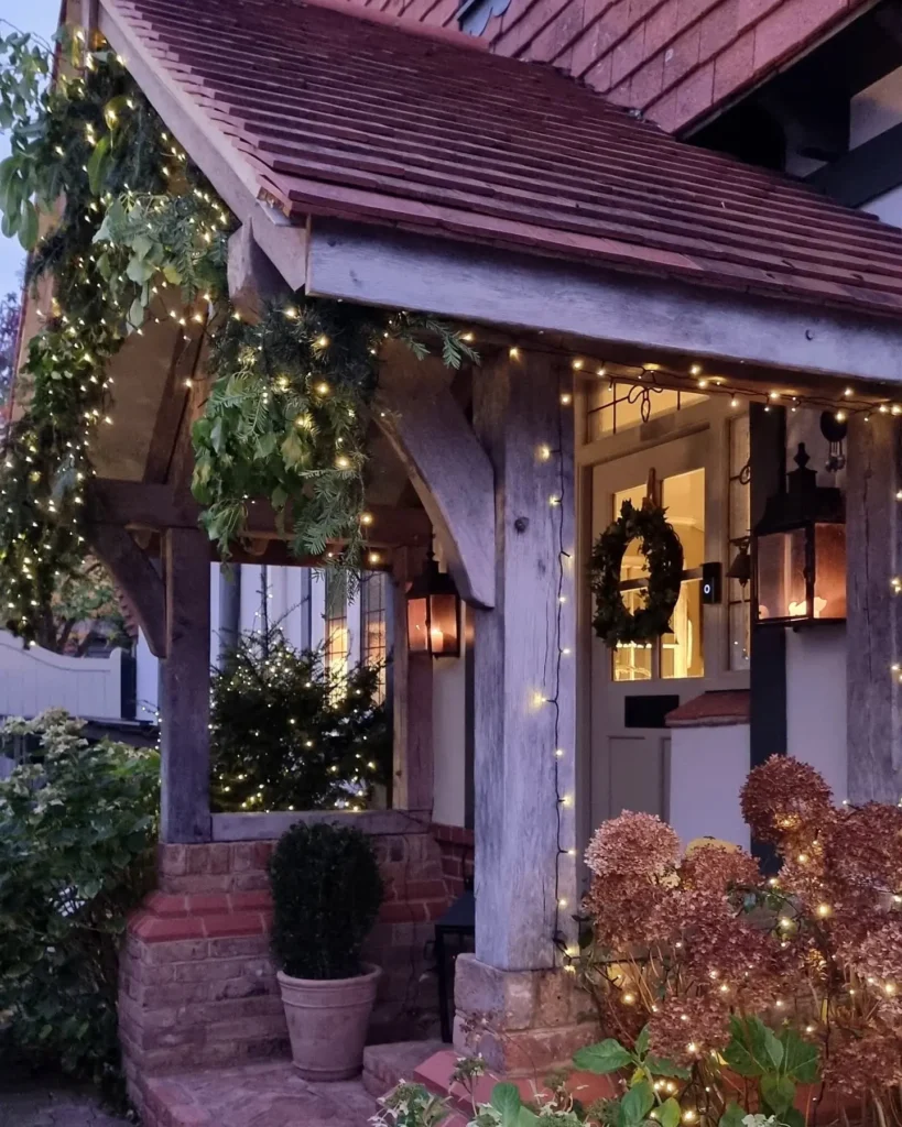 A front porch decorated with string lights, green garlands, and wreaths; potted plants and dried hydrangeas are visible by the entrance.