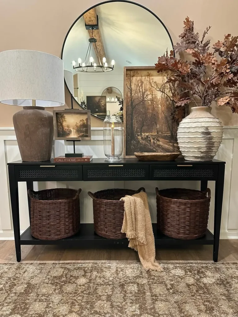 A console table with a lamp, framed art, a glass vase, and a ceramic vase with brown foliage stands in front of a round mirror, showcasing Thanksgiving entryway decor ideas; three woven baskets sit on the lower shelf.