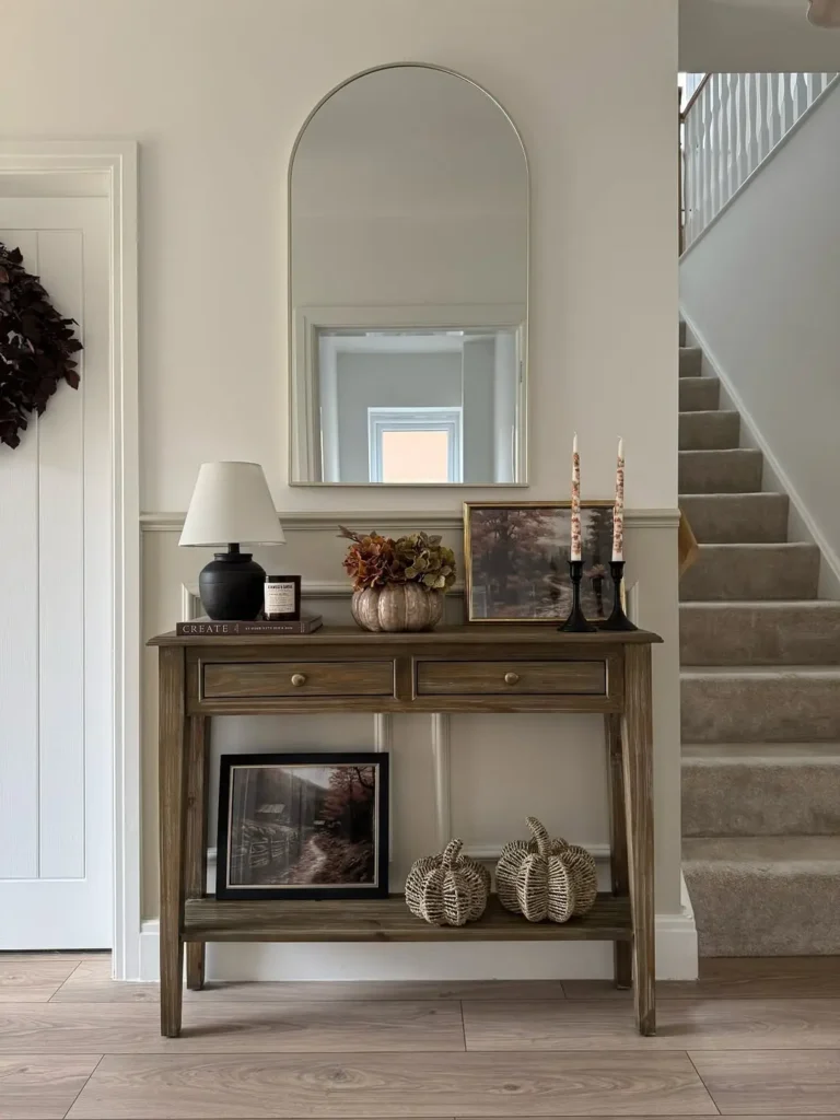 A wooden console table with decor items, including a lamp, framed pictures, candles, and wicker pumpkins, stands against a wall with a large arched mirror above it near a staircase, perfect for fall and thanksgiving entryway styling.