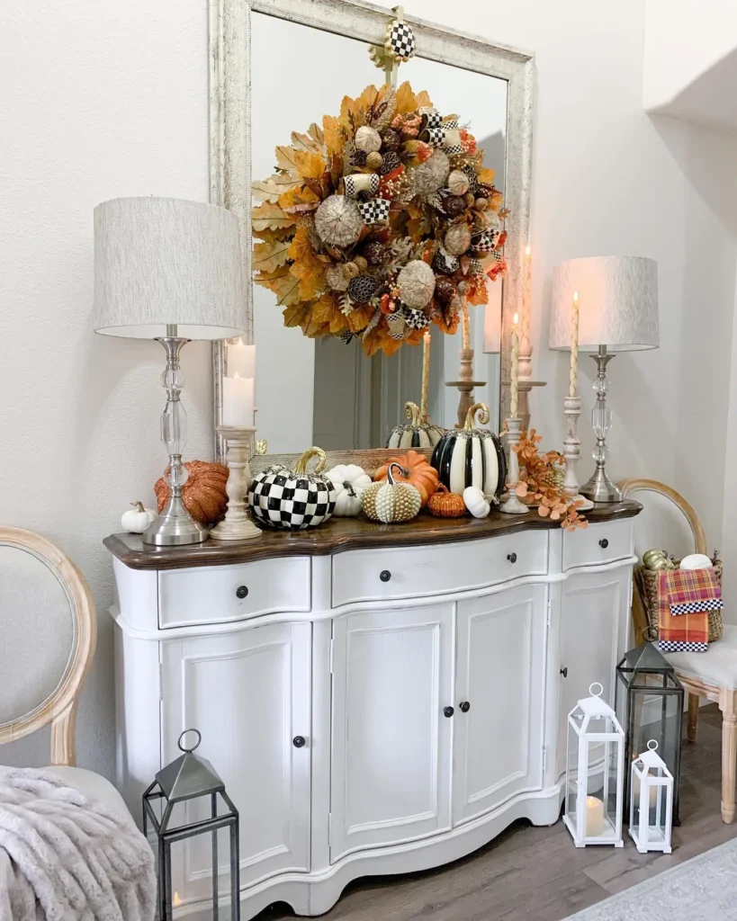 White sideboard with autumn-themed decor, including pumpkins, candles, and a leaf wreath on a large mirror; a throw blanket, two lamps, and lanterns add Thanksgiving entryway decor using light and texture to the inviting display.
