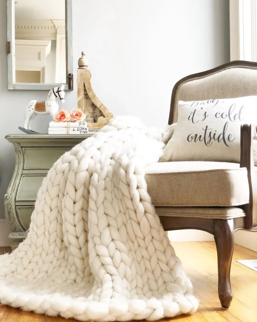 A beige armchair styled for winter with a chunky knit cream blanket draped over it, next to a side table with decor and a pillow that reads, "it's cold outside.