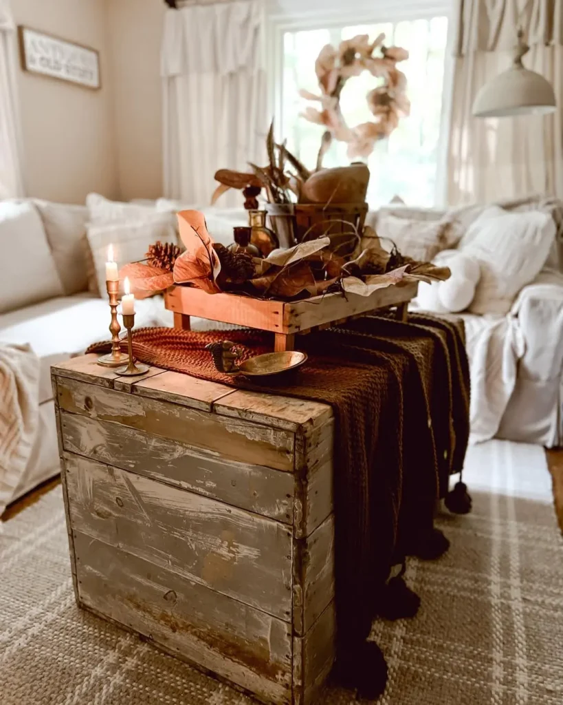 A rustic wooden trunk serves as a coffee table, topped with a brown throw, candle, and farmhouse fall decor in a cozy living room with white sofas and soft natural light.