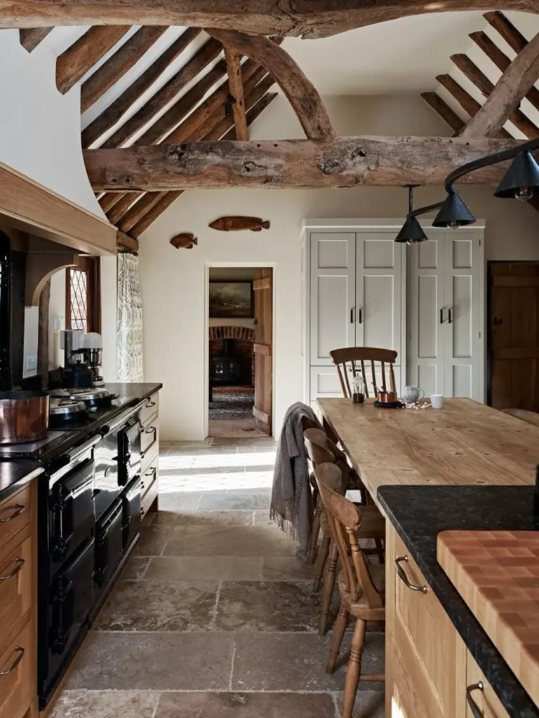 A functional and stylish rustic kitchen featuring exposed wooden ceiling beams, a large island with wooden chairs, stone flooring, and a black range cooker.