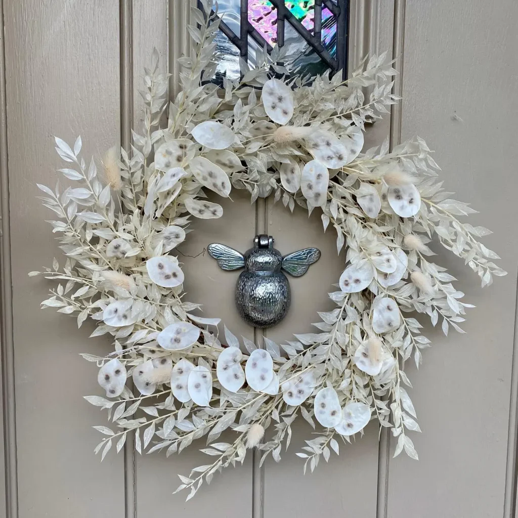 A winter wreath of pale, dried leaves and seed pods hangs on a beige door, centered around a metallic door knocker shaped like a bee with outstretched wings.