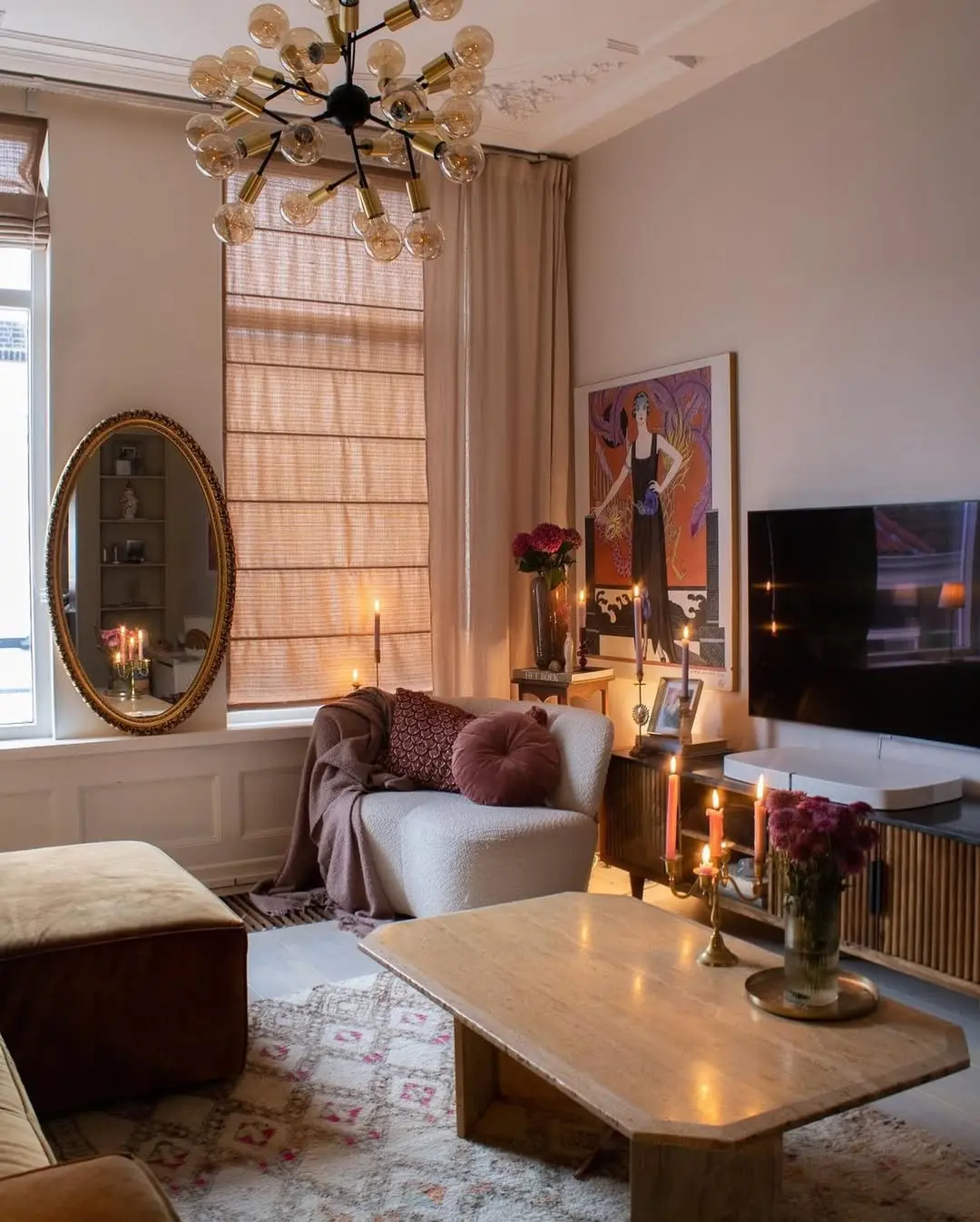 A cozy living room with a plush armchair, coffee table, sofa, wall art, candles, a TV, round mirror, and a window with light brown blinds. Warm lighting creates an inviting atmosphere, ideal for cozy winter nights.