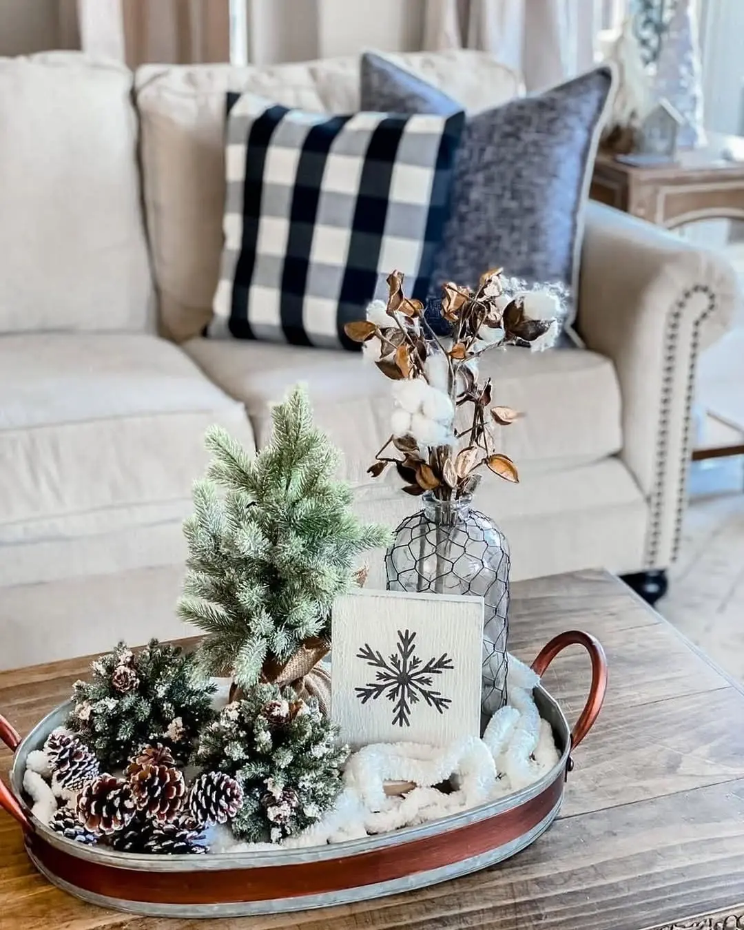 Winter decor vignette featuring a decorative tray with faux pine trees, pinecones, and a snowflake sign sits on a wooden coffee table in front of a beige sofa with plaid and velvet pillows.