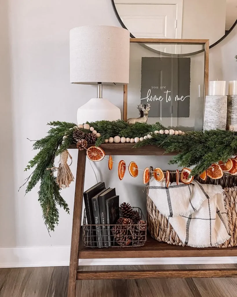 A wooden console table styled with winter entryway decor, pine garland, dried orange slices, books, pinecones, a cozy basket with a blanket, and a lamp. Framed art and candles adorn the top shelf for a warm seasonal welcome.
