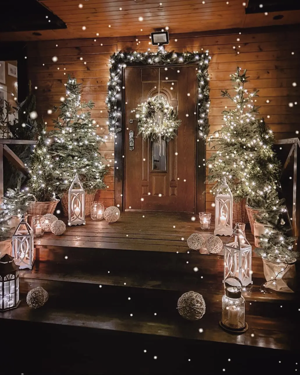 A wooden front porch winter scene, decorated with lit Christmas trees, lanterns, garlands, and a wreath on the door, as snow falls gently.