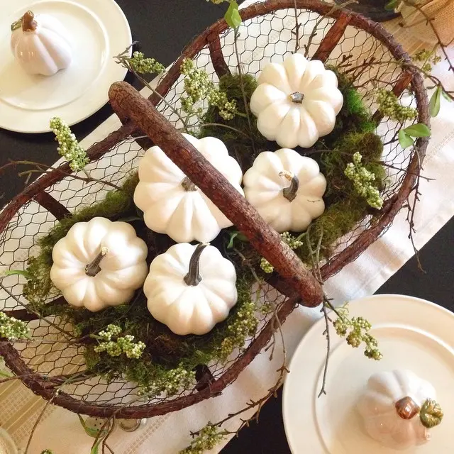 A rustic farmhouse basket filled with white decorative pumpkins and greenery sits as a centerpiece on a dining table set with plates and more white pumpkins.