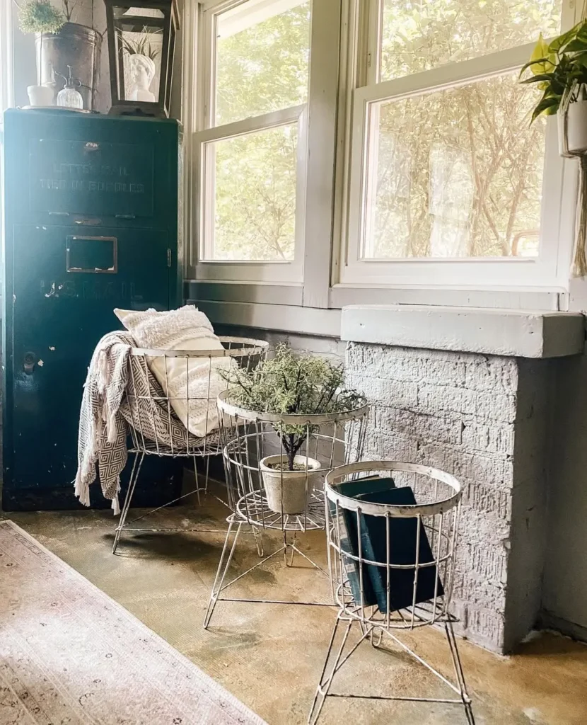 A cozy corner with wire-frame tables and chairs, a green metal cabinet, potted plants, and a stylish wire basket grouping, all bathed in light streaming through large windows.