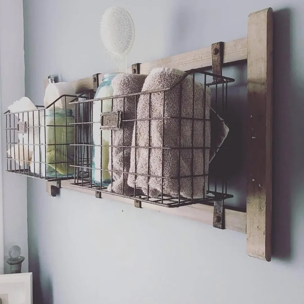 A wall-mounted farmhouse wooden and wire basket rack holds towels, bottles, and a brush against a light gray wall.