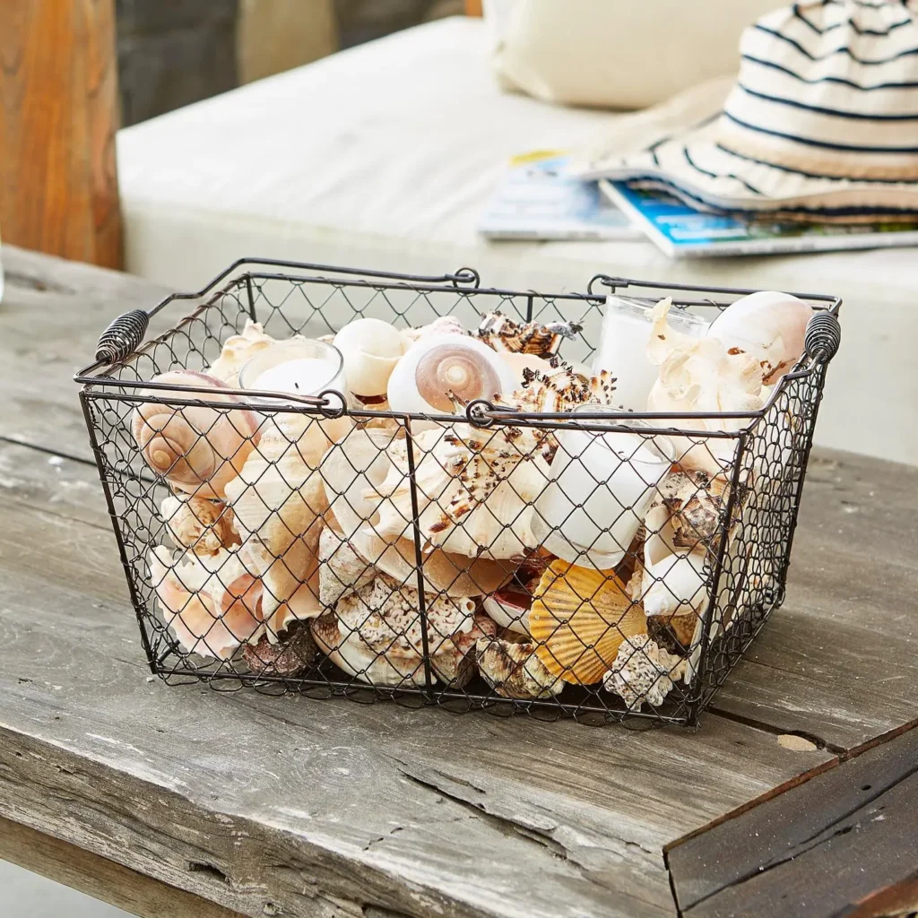 A wire basket filled with various seashells sits on a wooden table, adding farmhouse charm, with a striped bag and white cushion in the background.