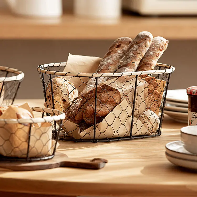 A wire basket filled with assorted bread, including baguettes and rolls, sits on a wooden table next to dishes and a jar, creating a cozy farmhouse vibe.