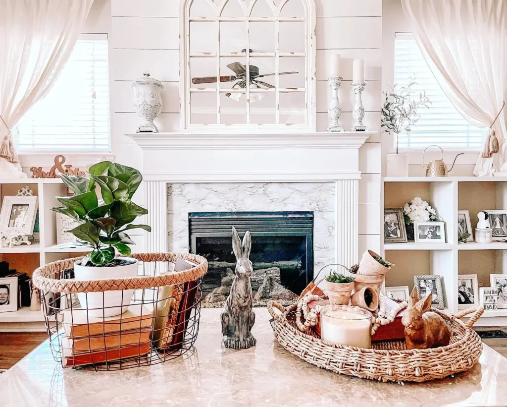 A living room with a marble fireplace, two bookshelves, and decorative items including plants, a wire basket, a candle, and ceramic rabbits on the coffee table to complete the farmhouse decor.