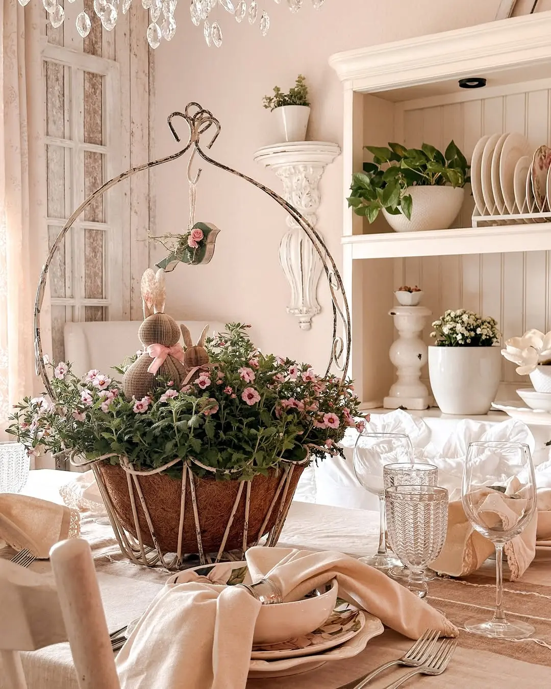 A dining table is set with glassware and neutral linens, featuring wire basket decor as a floral centerpiece with a stuffed bunny. Shelves in the background display plants and dishes.
