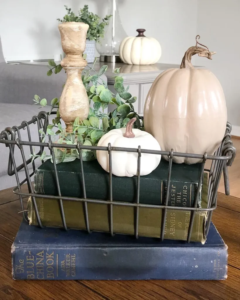 A metal basket with two decorative pumpkins, a wooden candle holder, faux greenery, and two books sits on top of a large blue book on a wooden table.