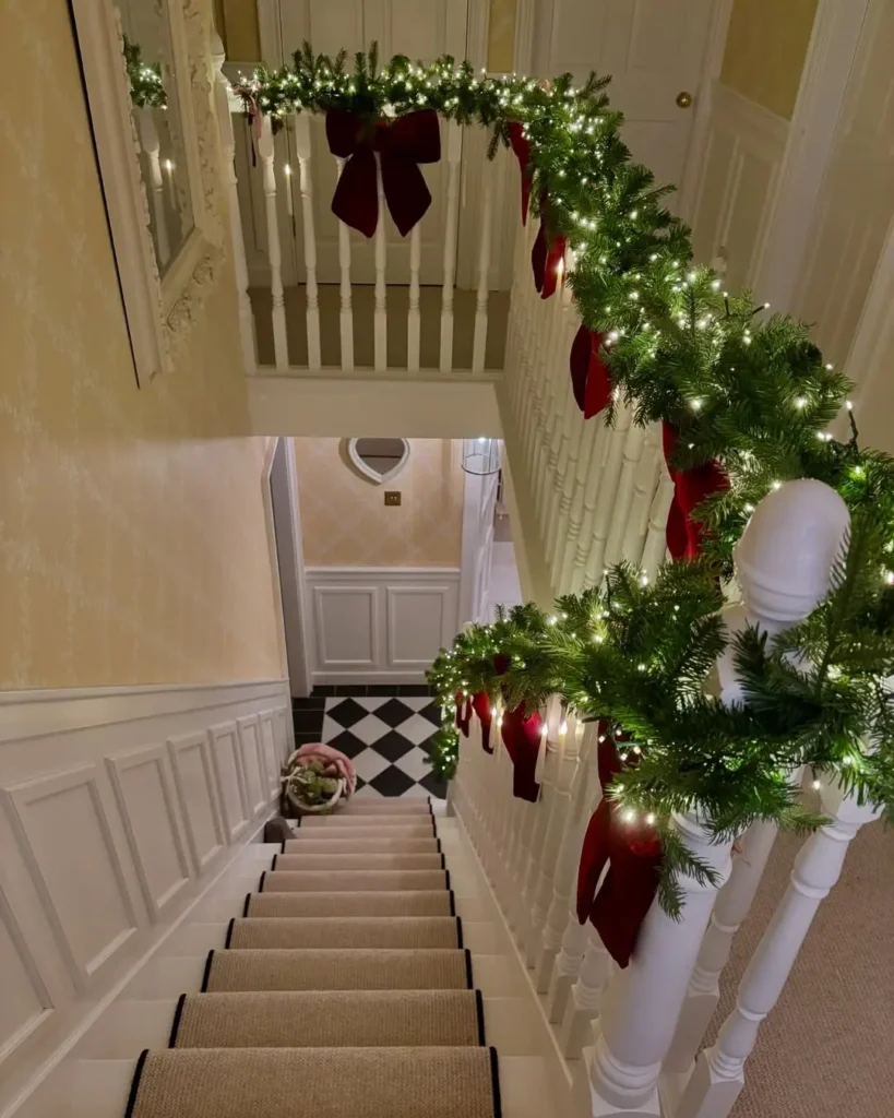 A staircase decorated with garland, red bows, and string lights, viewed from above; there is a checkered floor and a basket at the bottom of the stairs.