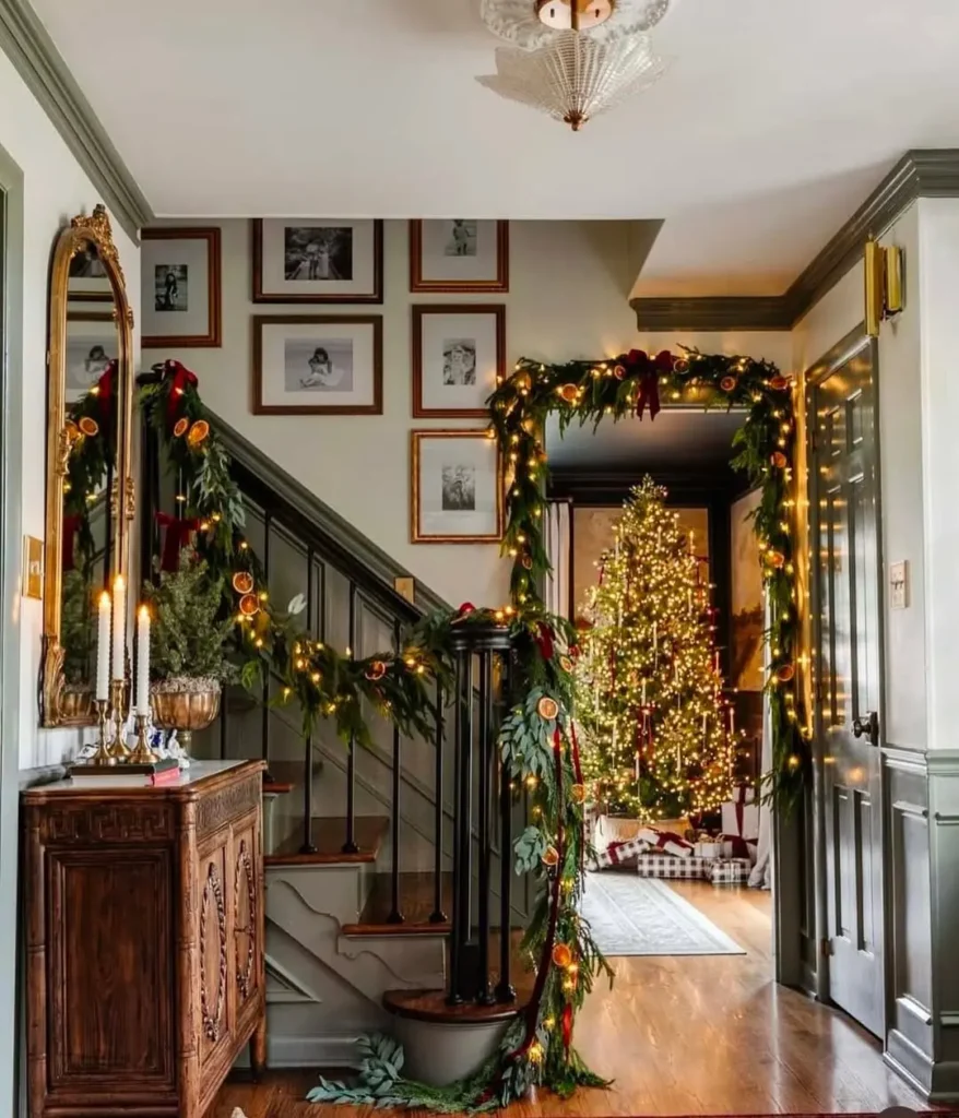 A hallway decorated for Christmas with garlands and lights, a mirror and candles on a wooden console, and a lit Christmas tree visible in the next room.