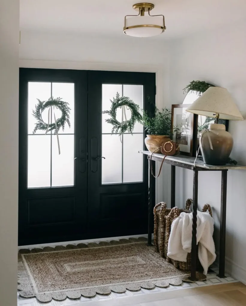 A tidy entryway styled for Christmas showcasing double black doors decorated with wreaths, a console table with a lamp and decor, and woven baskets holding blankets on a patterned rug.
