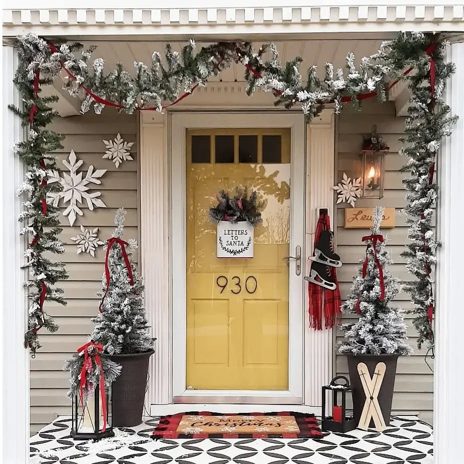 Easy Christmas Front Porch Decor Ideas come to life with garlands, artificial snow, red ribbons, snowflakes, small pine trees, a sled, lanterns, and a yellow door numbered 930 for a festive holiday welcome.