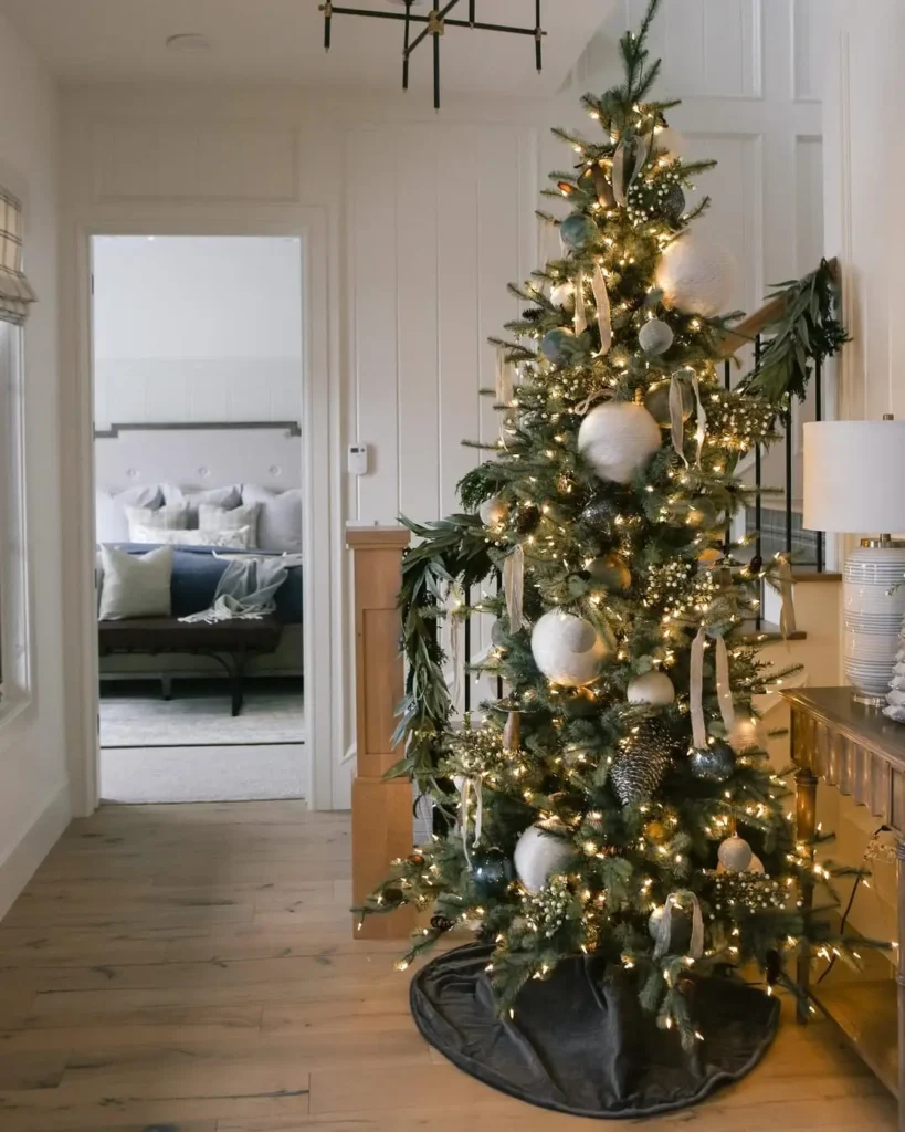 A decorated Christmas tree with white and silver ornaments and string lights stands in a hallway near a console table; a bedroom is visible in the background.
