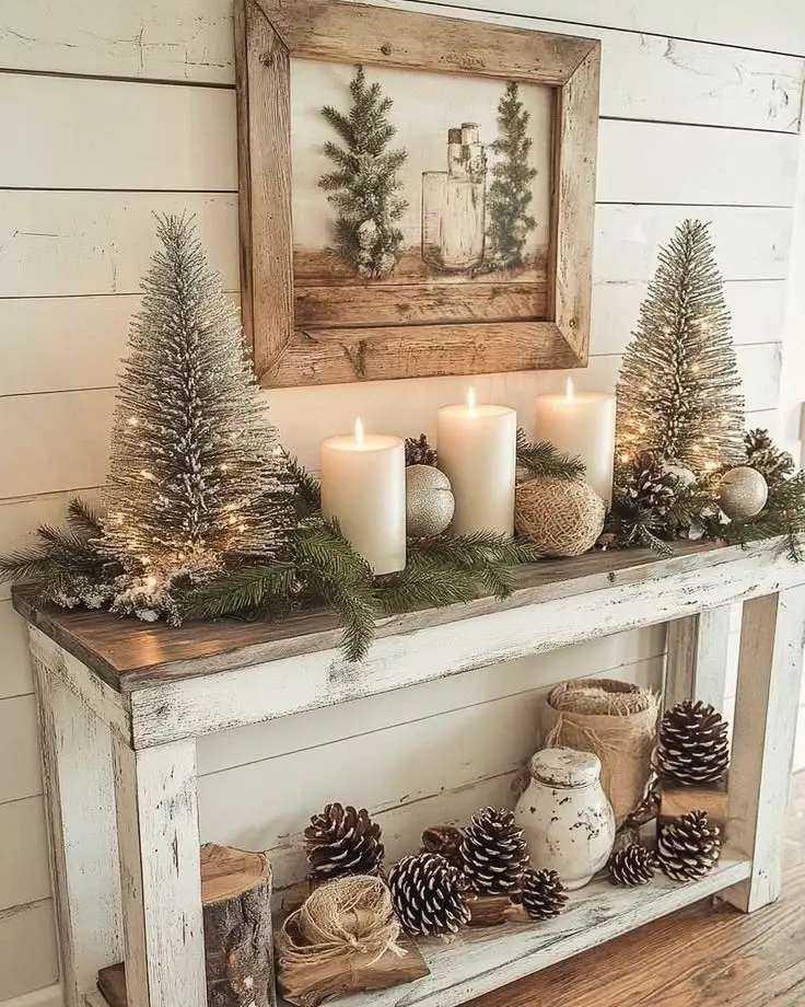 A rustic wooden console table creates a cozy Farmhouse Christmas Entryway, adorned with snowy artificial trees, white candles, pinecones, ornaments, greenery, and a wintery framed art piece above.