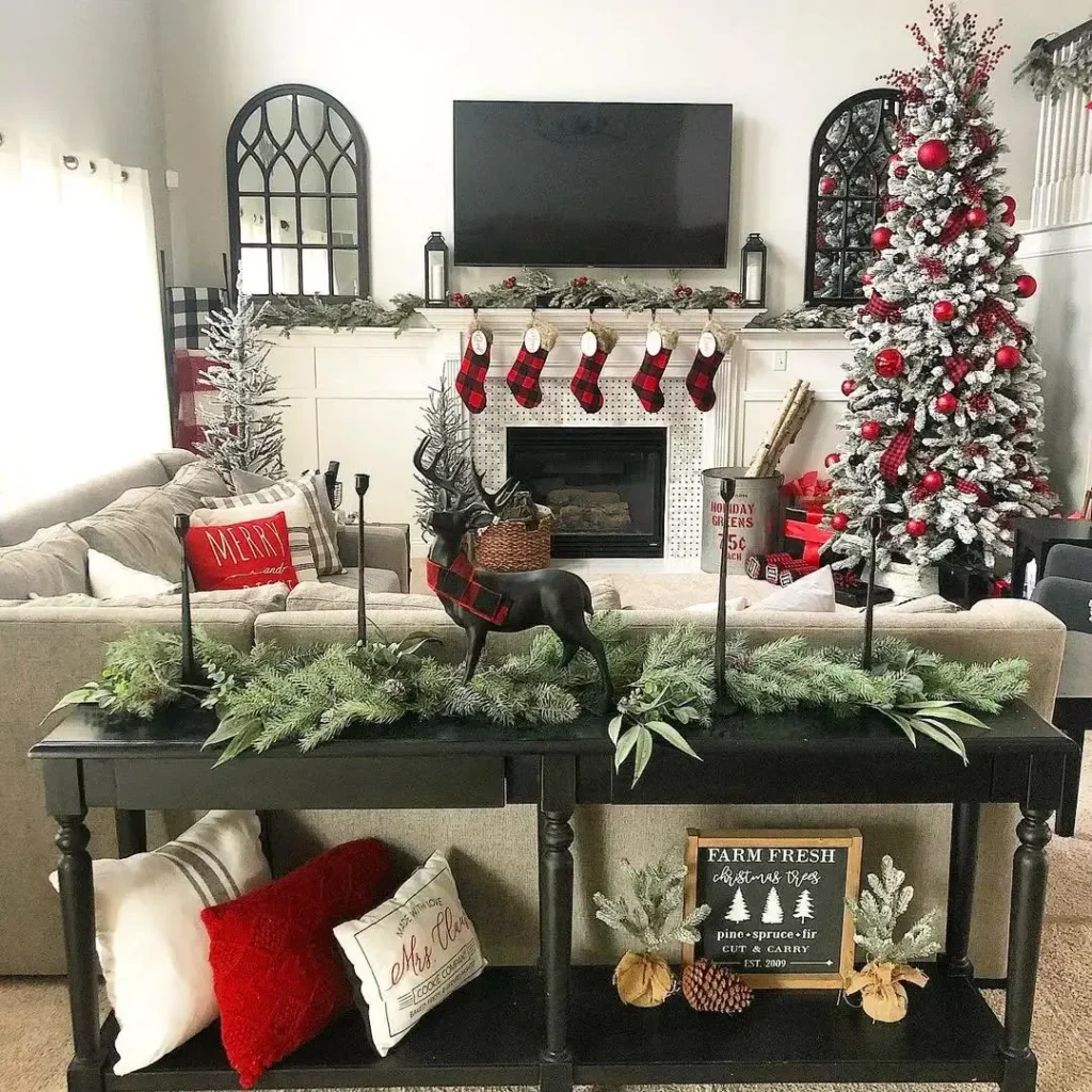 Living room decorated for Christmas with a tree, stockings above the fireplace, red and white accents, festive greenery on a black console table, and cozy farmhouse pillow covers adding rustic charm.