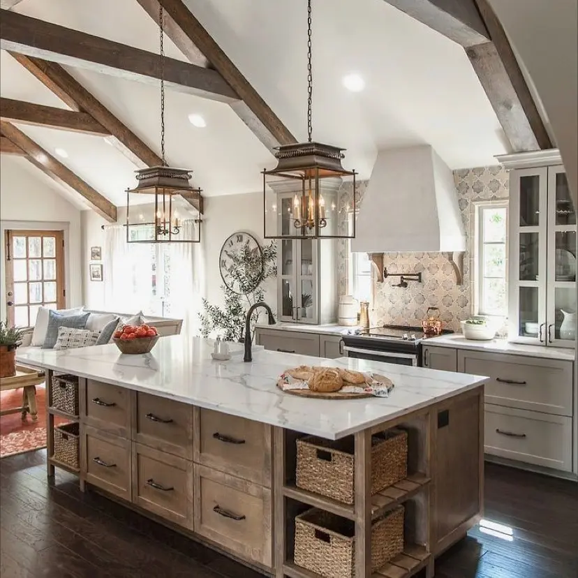 Bright French country farmhouse kitchen with exposed wooden beams, large marble island, pendant lights, wicker baskets, white cabinetry, and stove with decorative backsplash.