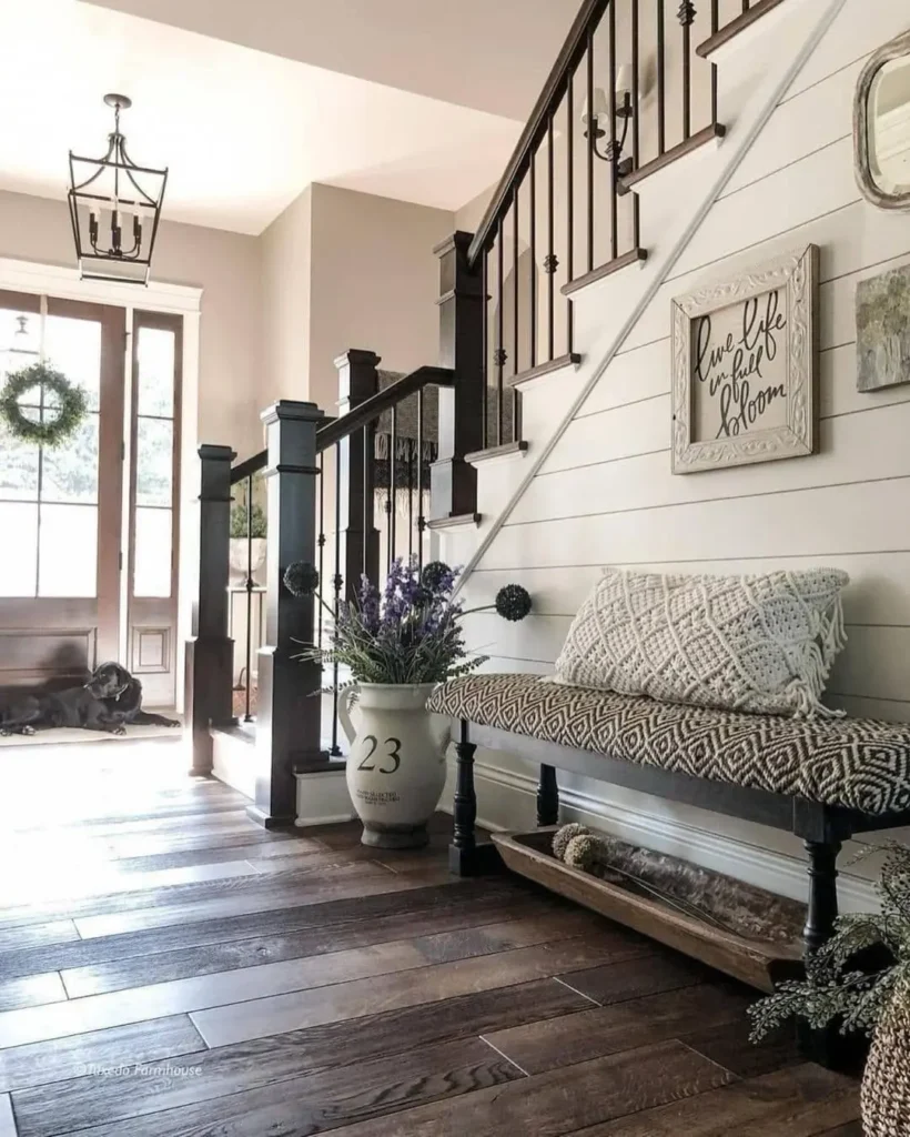 A bright entryway with wood floors features a bench with cushions, a large vase of flowers, a staircase with black metal railings, and wall art above the bench.