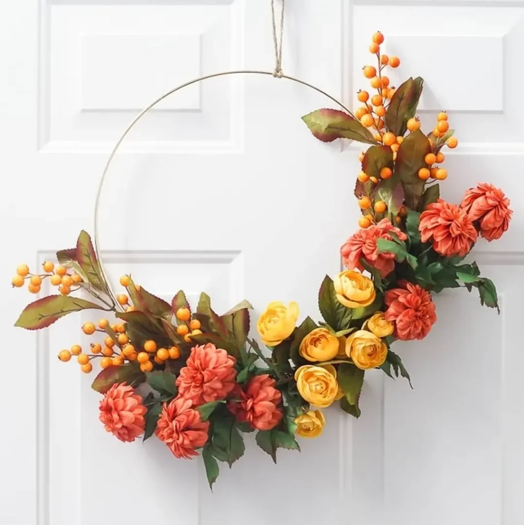 A modern minimalist fall wreath with orange and yellow flowers, green leaves, and orange berries, hanging on a white door.