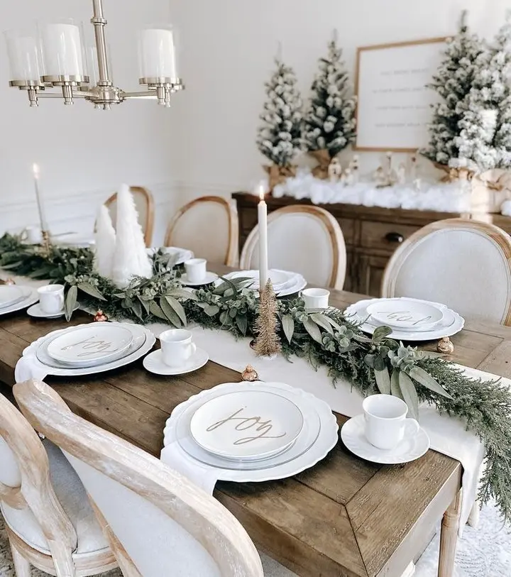 A christmas dining table is set with white plates that have "joy" written on them, greenery garland, candles, and festive decor. Snow-covered artificial trees are displayed on a sideboard in the background.