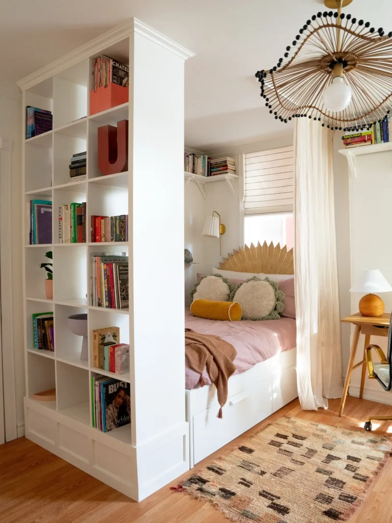 A cozy studio apartment using furniture to separate zones, with a bed tucked behind a white bookshelf room divider, decorative pillows, a wooden desk, books, and a patterned rug on a wooden floor.