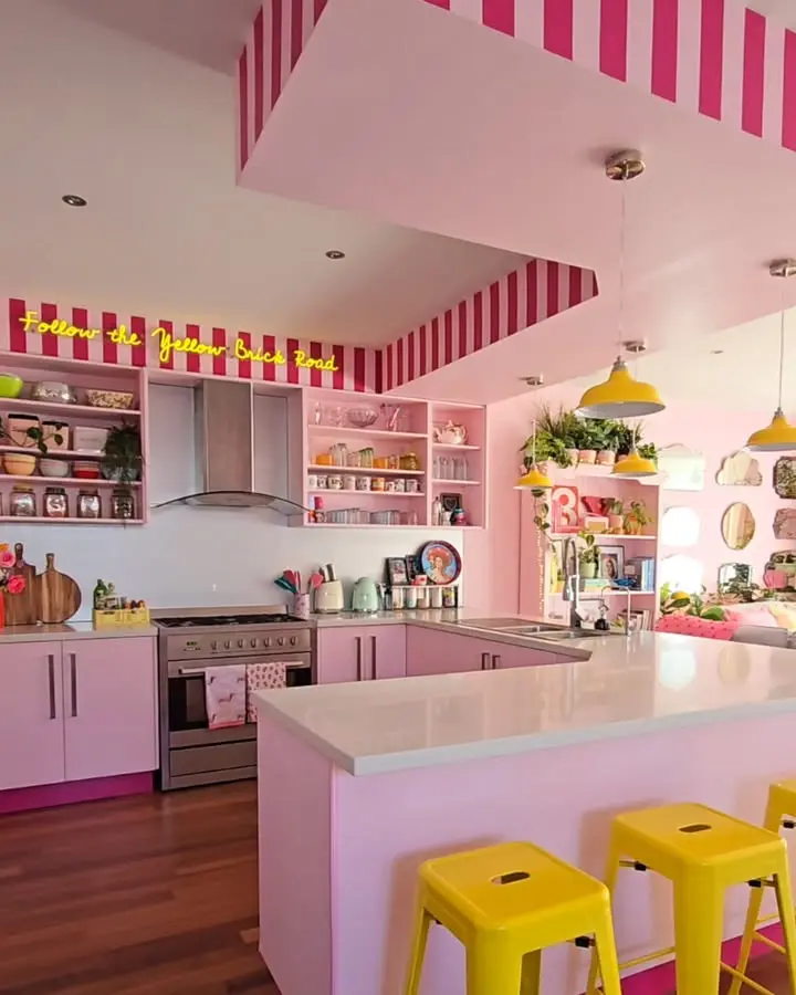 A vibrant pink kitchen with yellow stools, white countertops, open shelving, and striped accents. Neon sign above reads "Follow the Yellow Brick Road." Various kitchenware is visible.