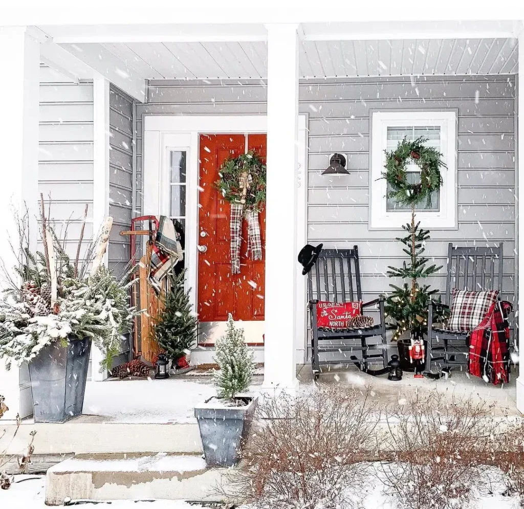 A festive christmas front porch features a snowy red door, holiday wreaths, small Christmas trees, two rocking chairs, a sled, and plaid blankets to complete the cozy scene.