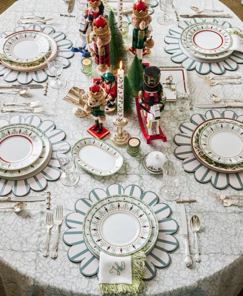 A round table set with holiday-themed plates, silverware, glasses, nutcracker figurines, small trees, and candles on a festive christmas table linen.