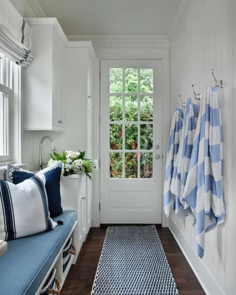 A coastal mudroom featuring a glass door, blue-and-white striped towels on hooks, a cushioned bench with pillows, a rug, and a window; greenery is visible outside.