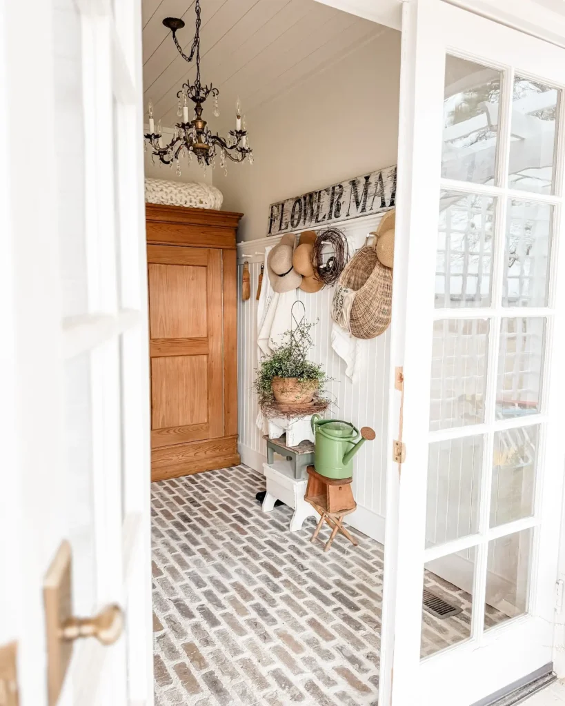 A small french country mudroom with brick flooring, a wooden wardrobe, hanging hats, a "FLOWER MARKET" sign, potted plants, and a green watering can next to a white-paneled wall.