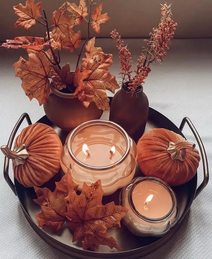 A tray displays two lit candles, artificial pumpkins, autumn leaves, and small vases with dried foliage, creating a fall-themed decor arrangement.